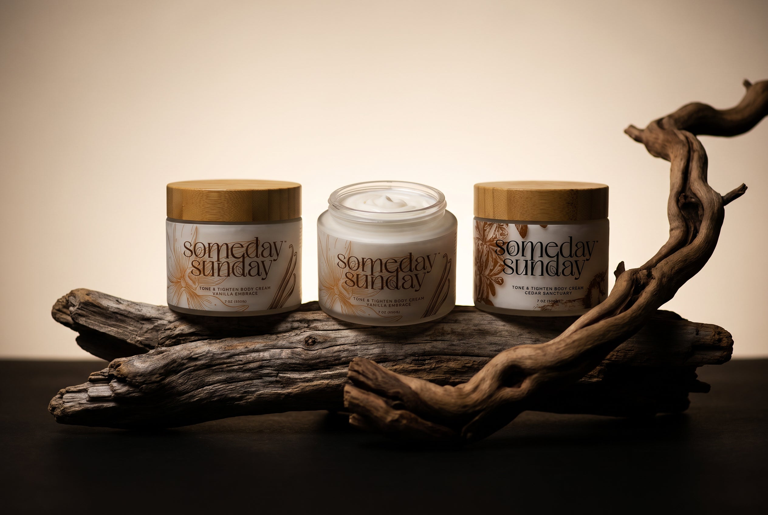 Three Someday Sunday body cream jars on weathered driftwood against a warm cream background, lit low and moody for fall.