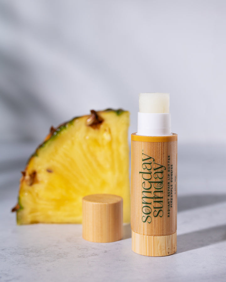 Someday Sunday Pineapple Sunrise lip butter stick in its bamboo case beside fresh pineapple.