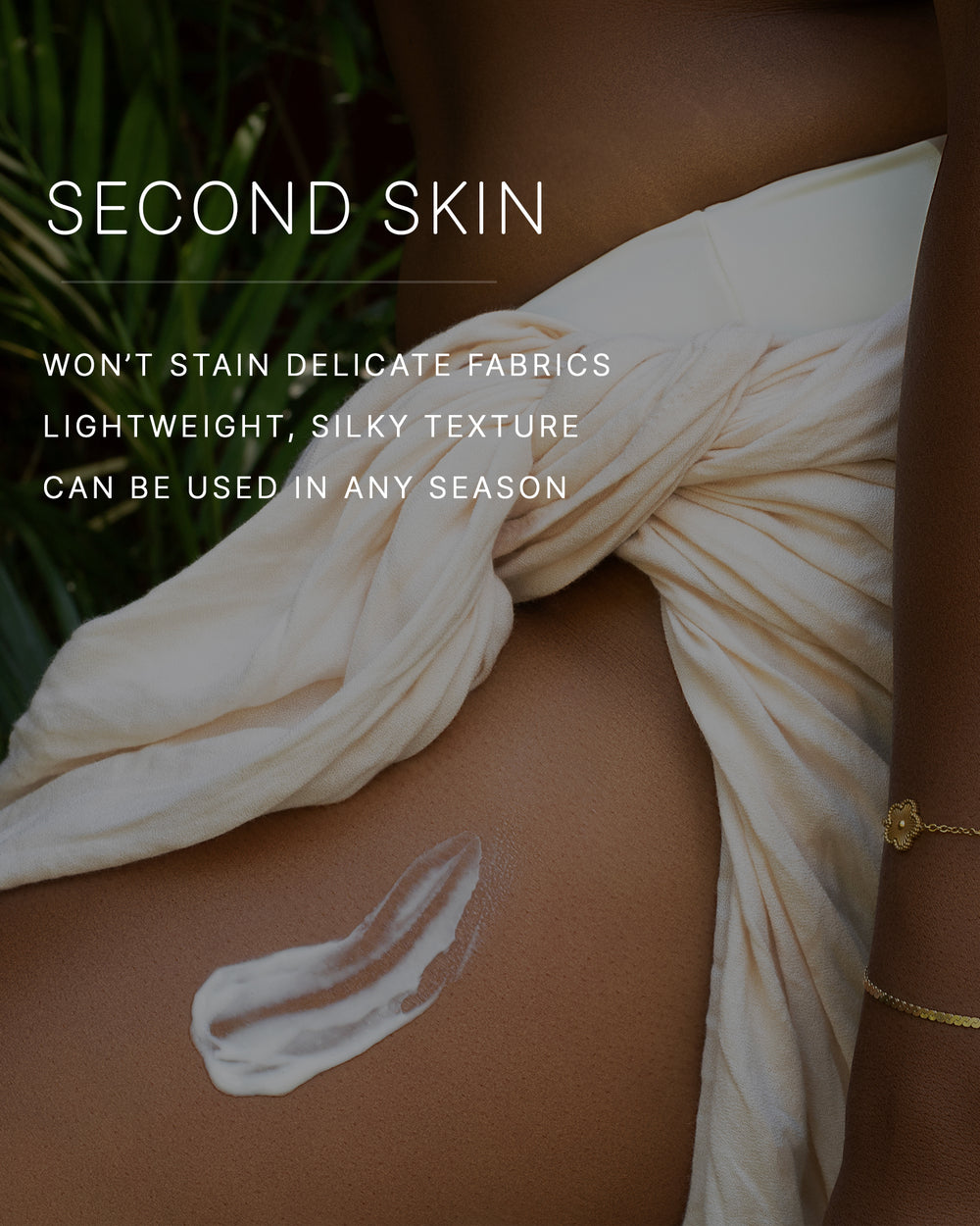 Second skin. Won't stain delicate fabrics. Lightweight, silky texture. Can be used in any season. A swipe of body cream smoothed onto the hip of a woman with a deep skin tone wearing a cream wrap, with tropical greenery behind her.
