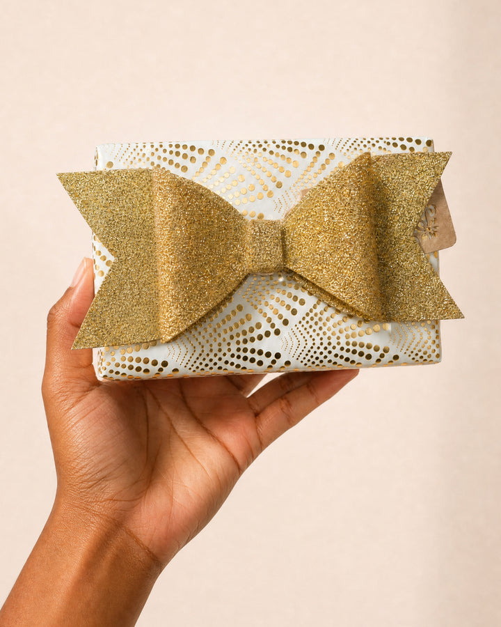 A hand holding a small gift wrapped in white and gold paper with a large gold glitter bow.