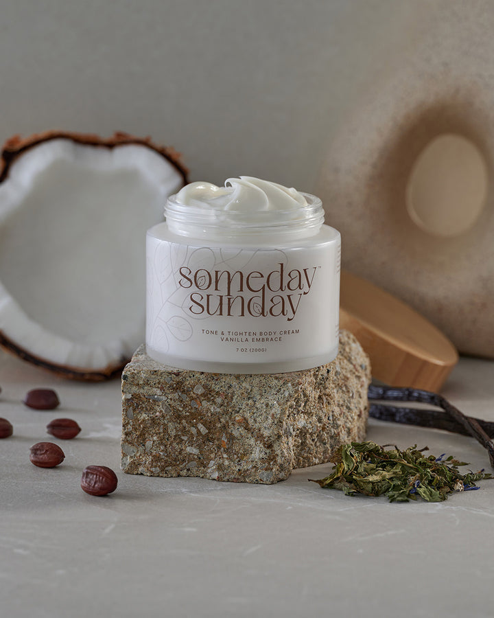 Someday Sunday Vanilla Embrace body cream on a stone plinth beside a halved coconut.