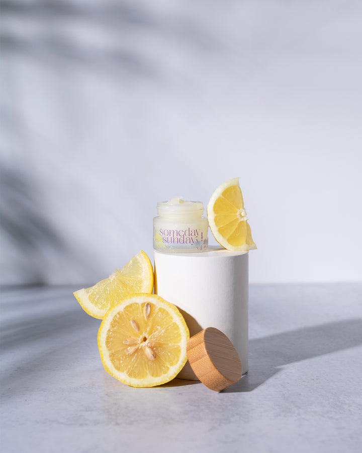 Someday Sunday Lavender Lemonade lip butter jar beside fresh lemon slices.