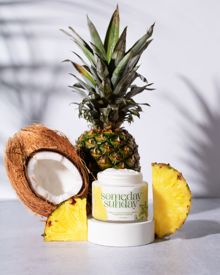 Someday Sunday Pineapple Sunrise body cream beside a fresh pineapple and halved coconut.
