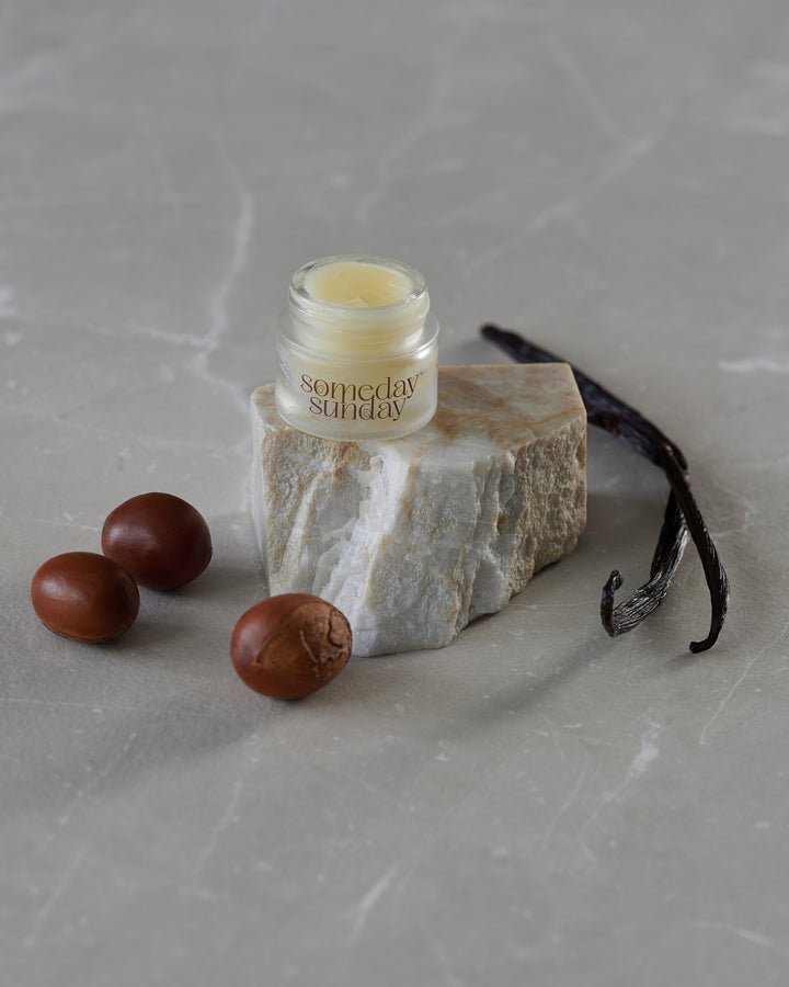 Someday Sunday Vanilla Embrace lip butter jar on stone with vanilla pods.