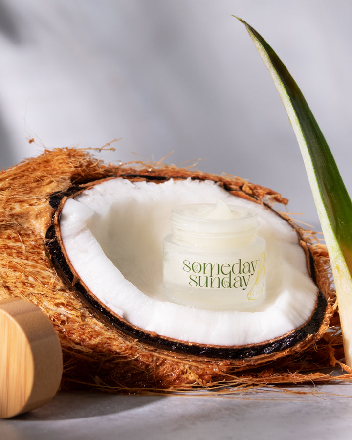 A Someday Sunday lip butter jar nestled in a halved coconut.